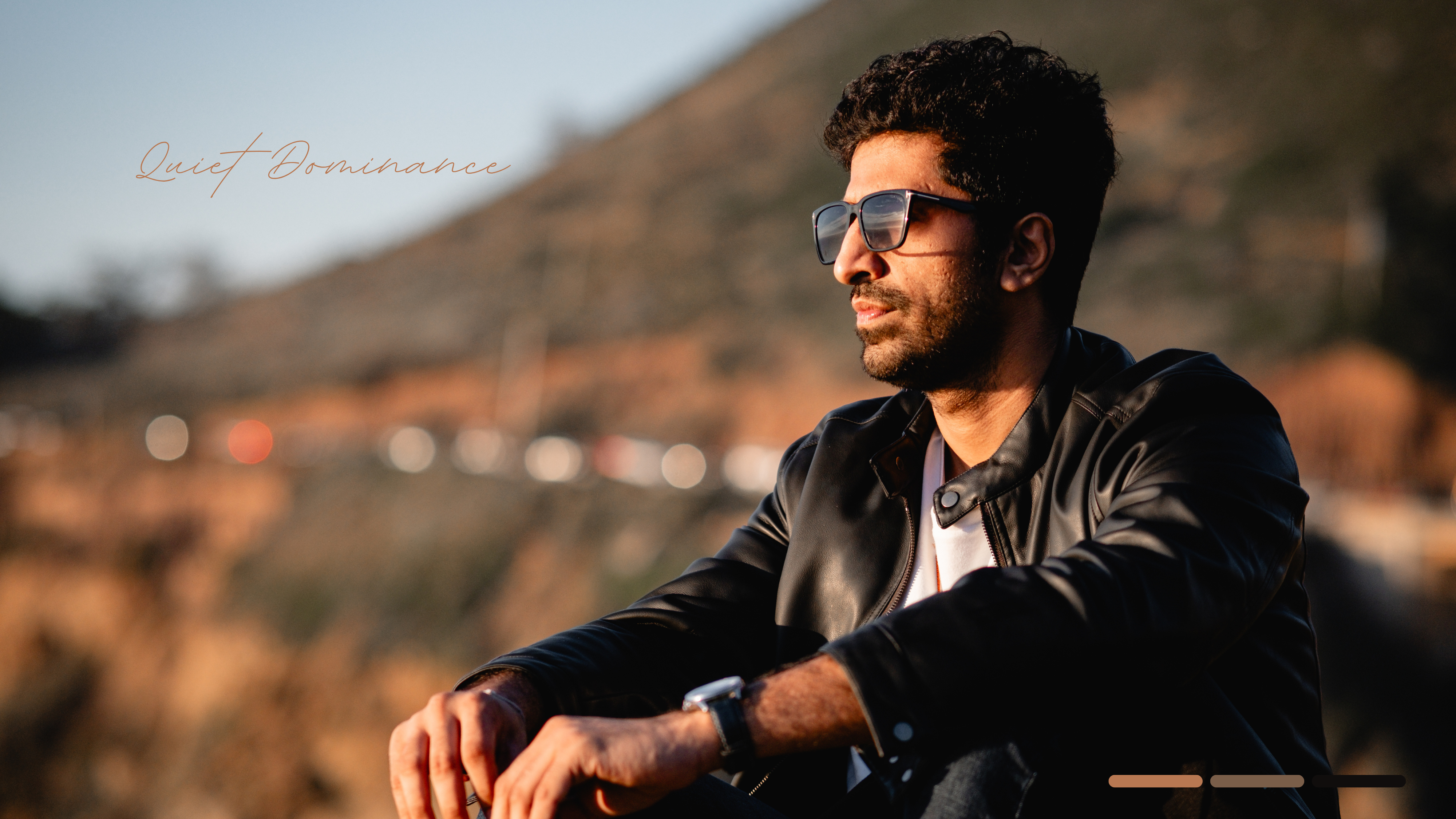 Man in leather jacket and sunglasses seated on a hillside at golden hour